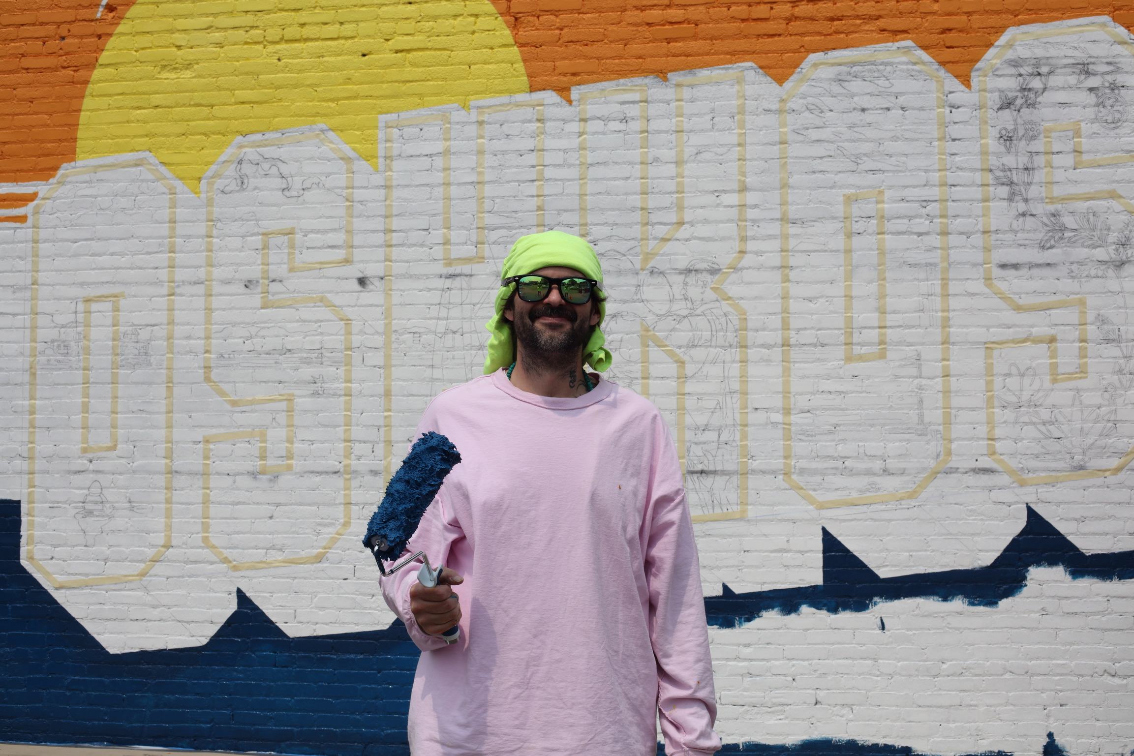 American Mural artist holding a paint roller in front of the outlined Greetings from Oshkosh mural.