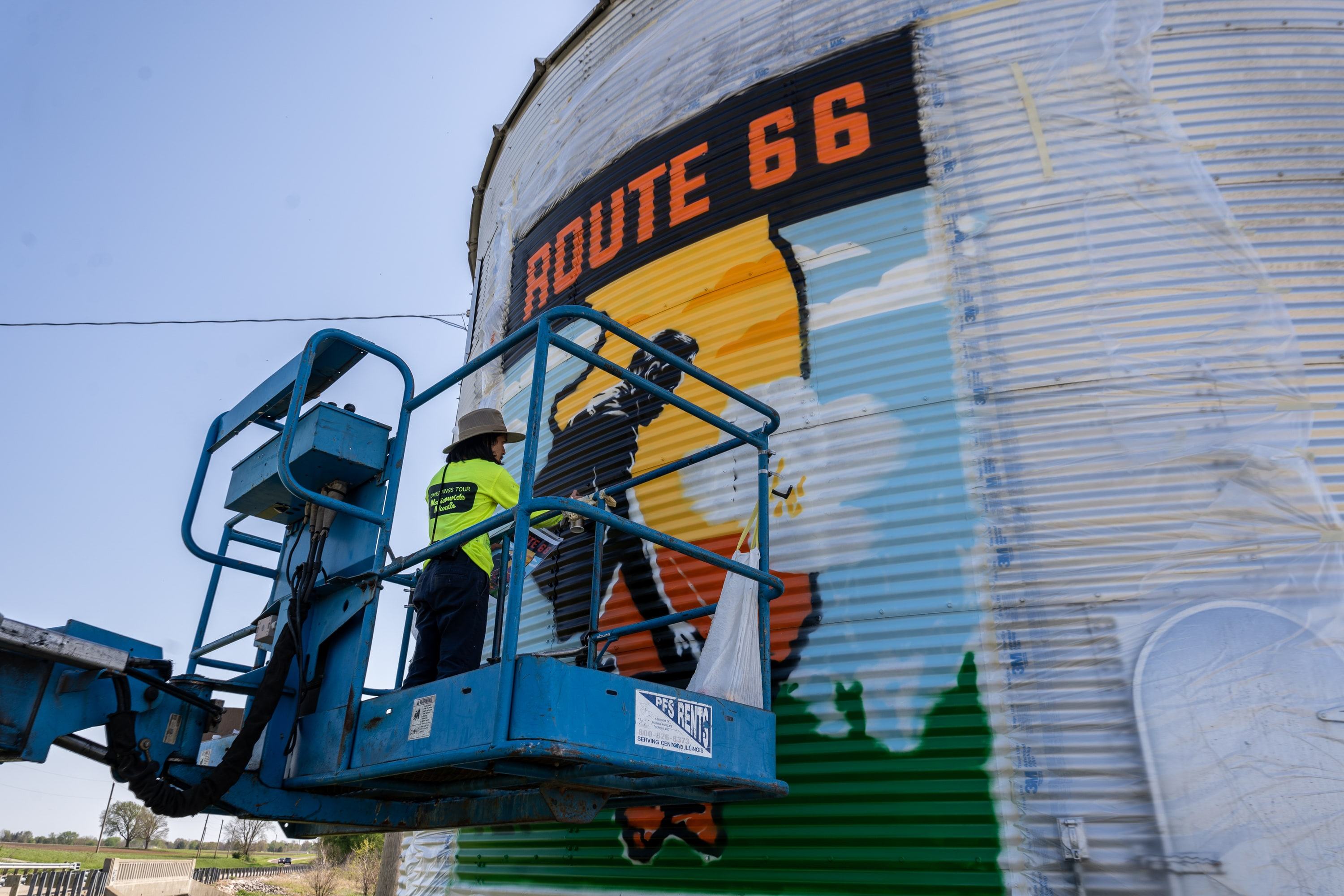 American Mural artist painting the Route 66 mural from a boom lift on a curved metal building.