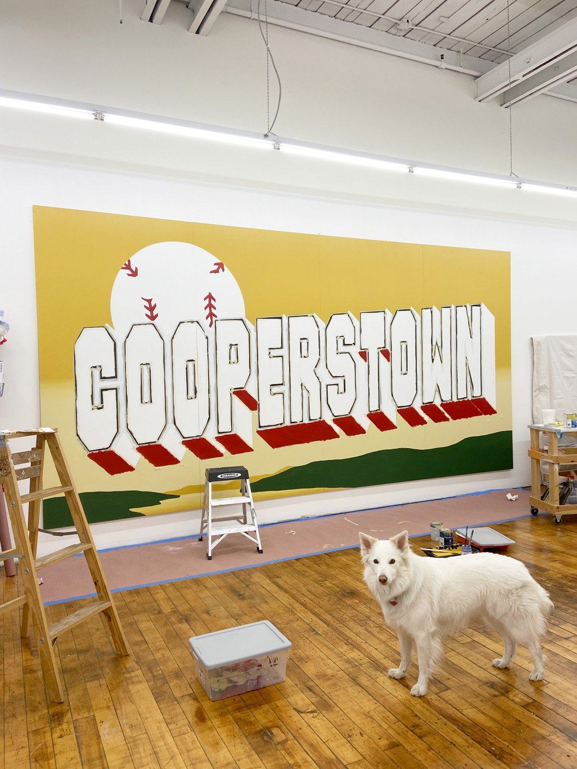 Partially painted Cooperstown mural staged in the studio with ladders, paint supplies, and a white dog.