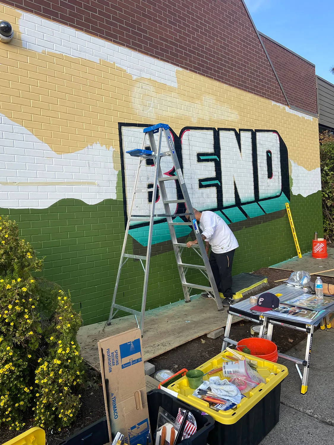 Artist painting the yellow background and letter outlines on the Bend mural