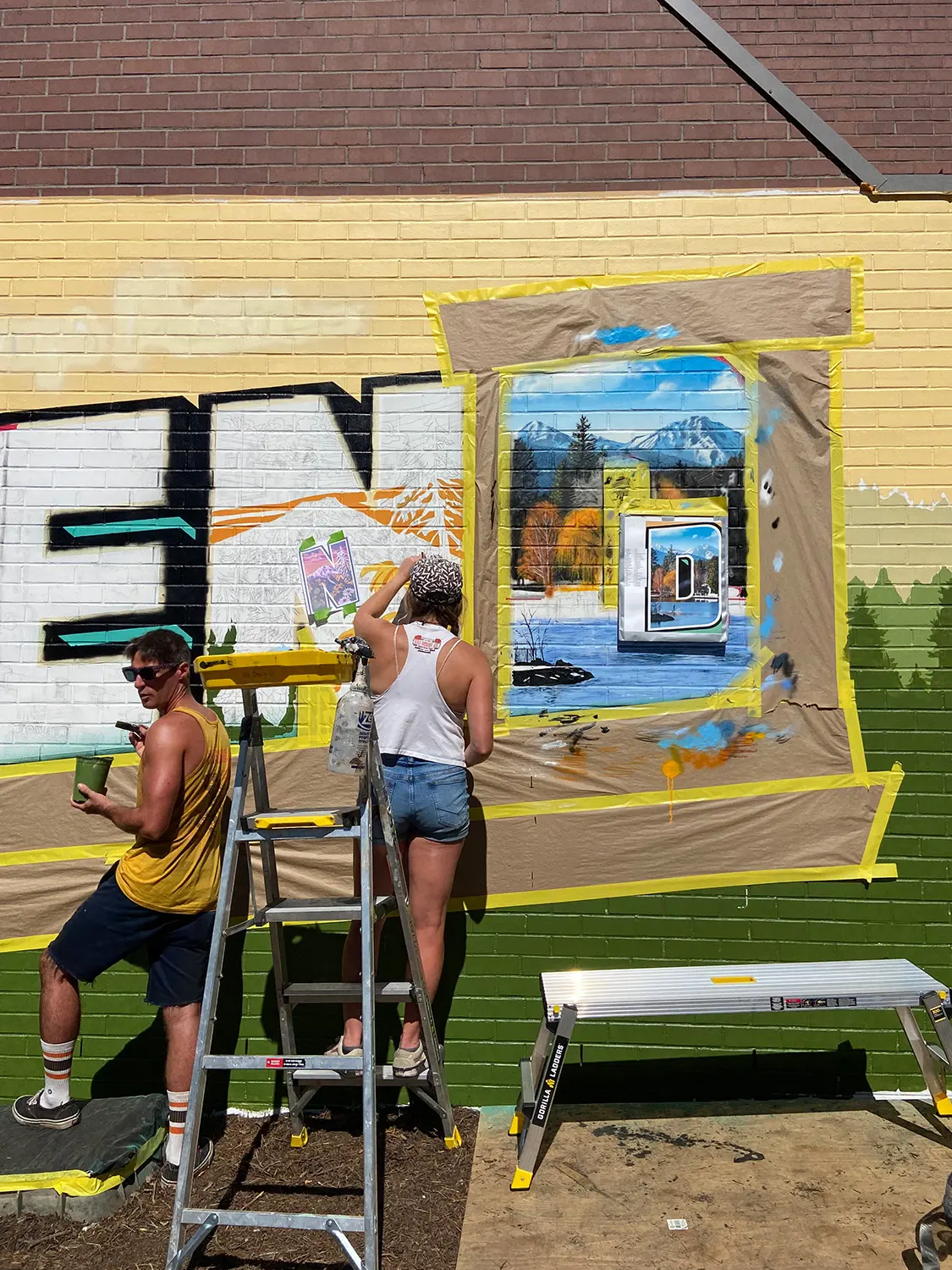 Two artists painting landscape details inside the Greetings from Bend mural lettering.