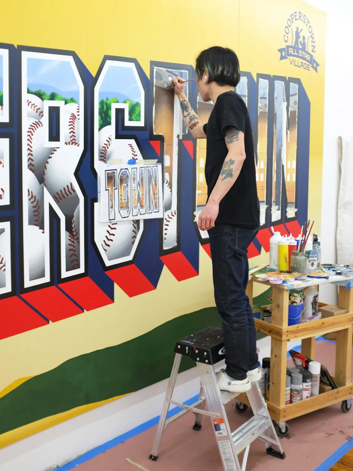 Artist finishing baseball details on the Cooperstown mural from a step stool