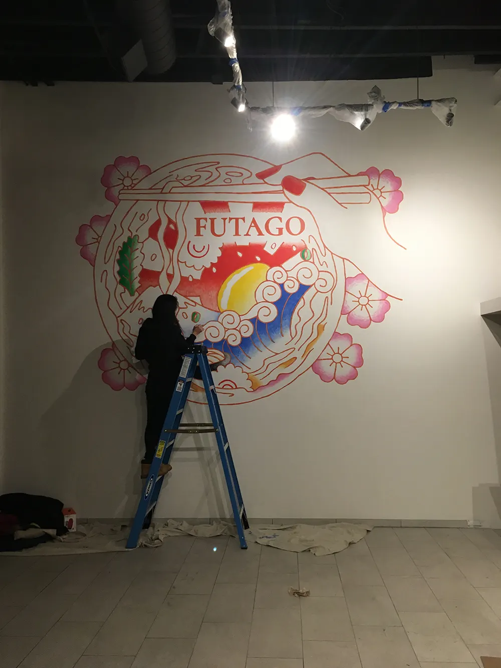 Artist working from a ladder on the udon bowl mural before the restaurant opened