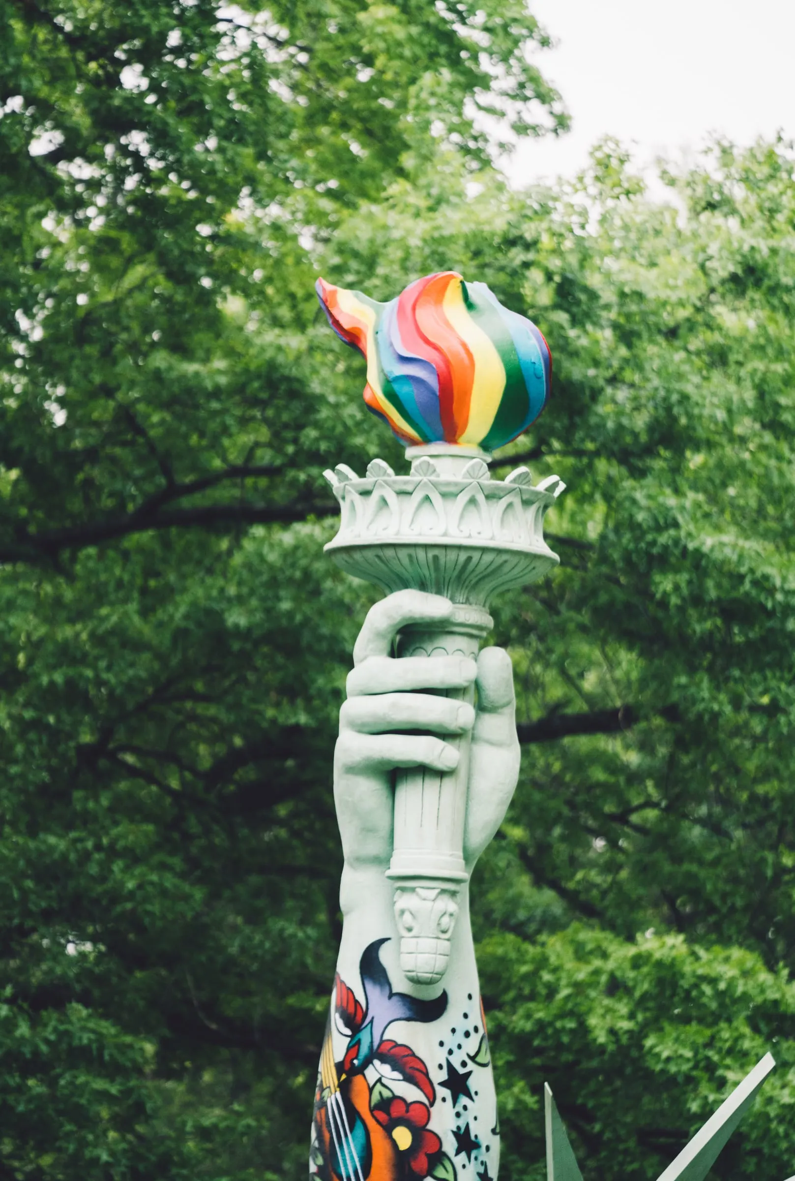 Close view of the rainbow-painted torch above a colorful bird and flower tattoo on the sculpture's arm