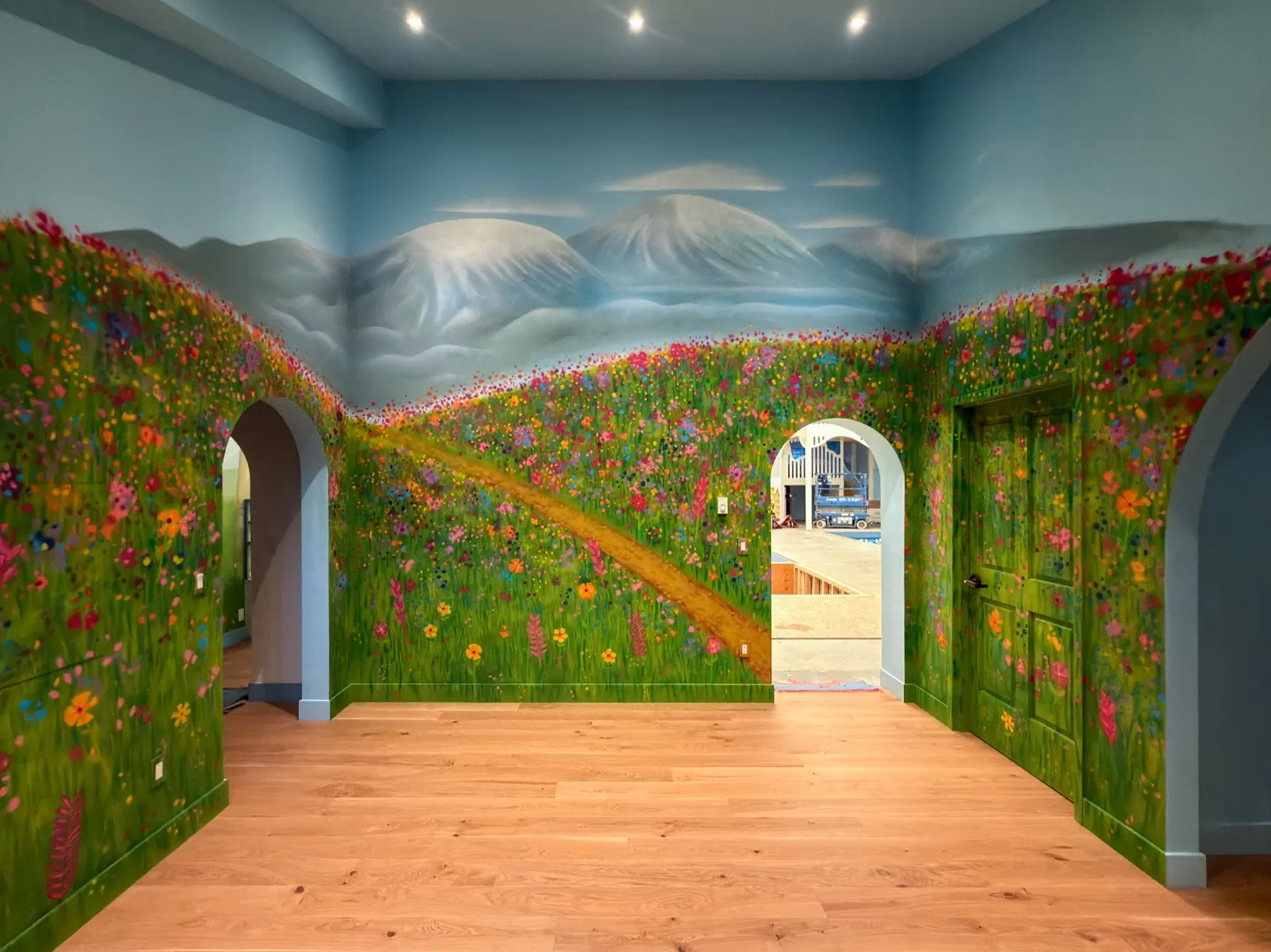 Wide view of the completed playroom mural with wildflower hills, two snowy mountain peaks, arched openings, and a painted green door