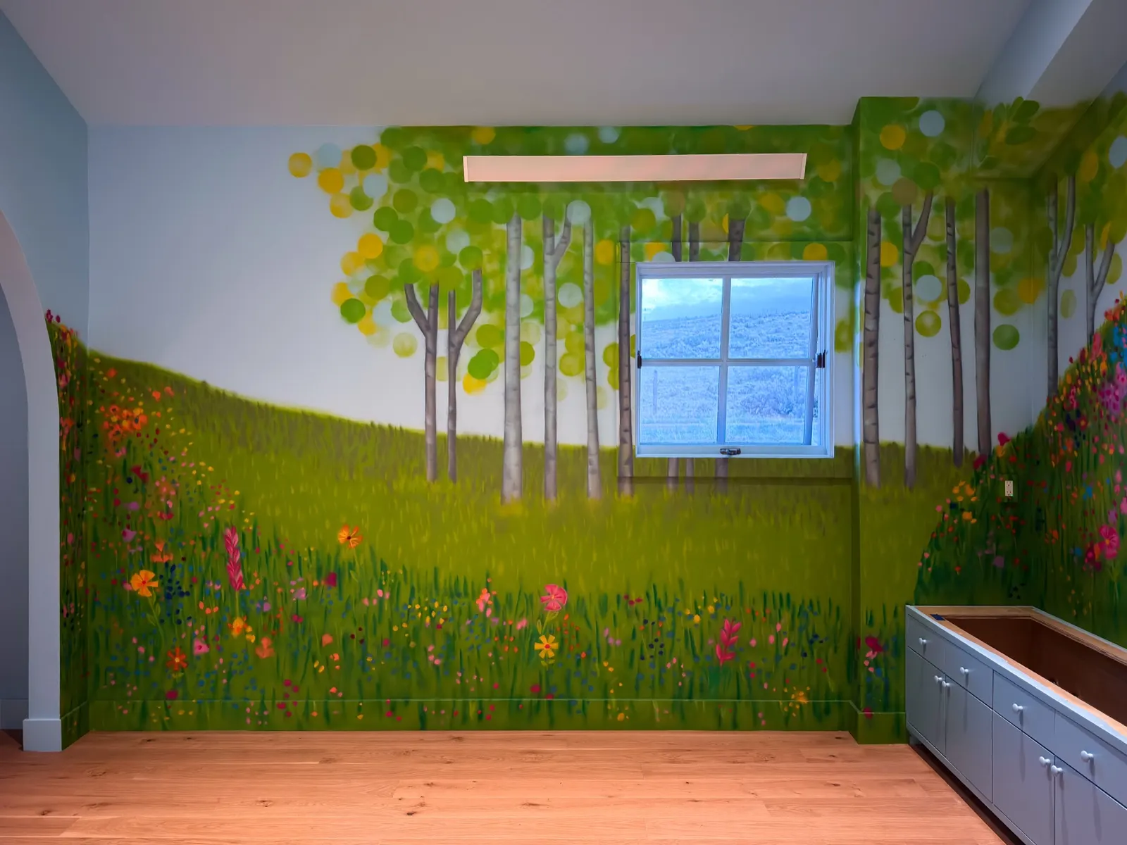 White-trunked trees and a grassy wildflower meadow painted around a small playroom window
