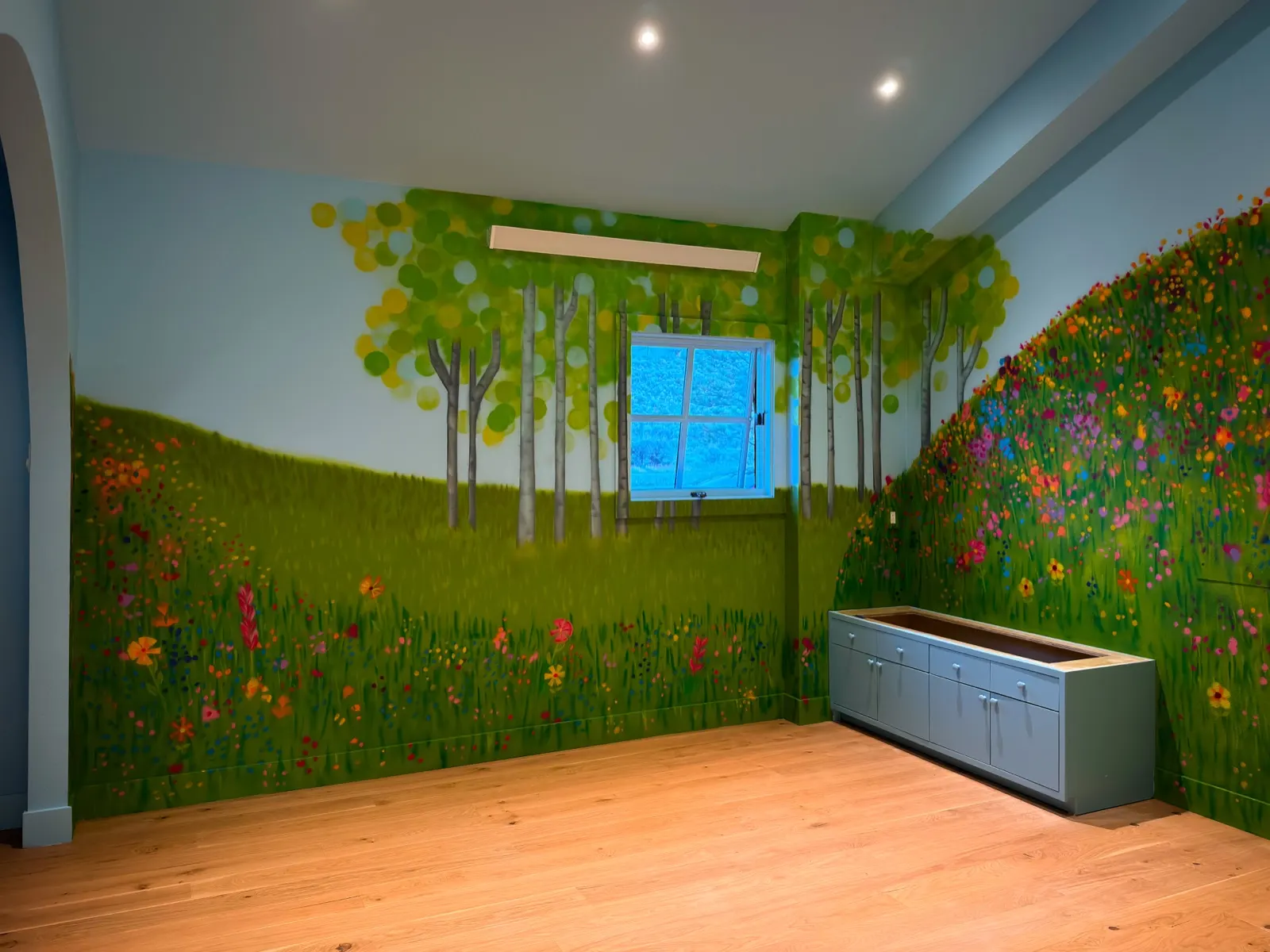 Wide playroom view with white-trunked trees, flowered hills, a window, and low gray cabinetry