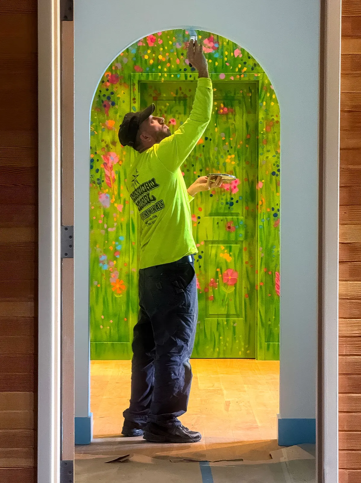 American Mural painter brushing a finishing detail onto the rounded opening, with the painted green door beyond
