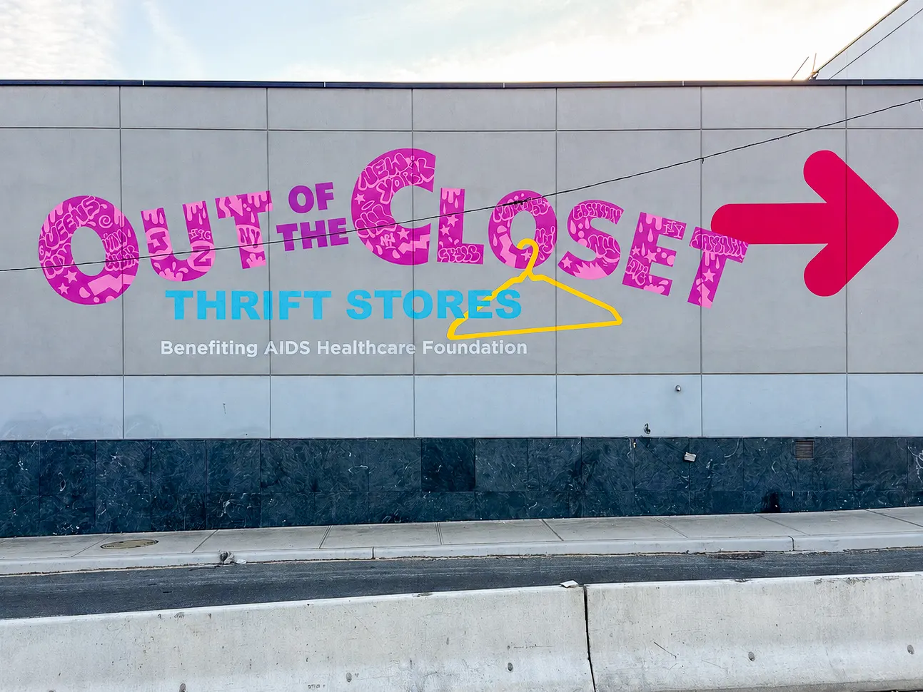 Wide exterior view of the completed Out of the Closet mural with pink lettering, turquoise Thrift Stores text, a yellow hanger, and a red arrow