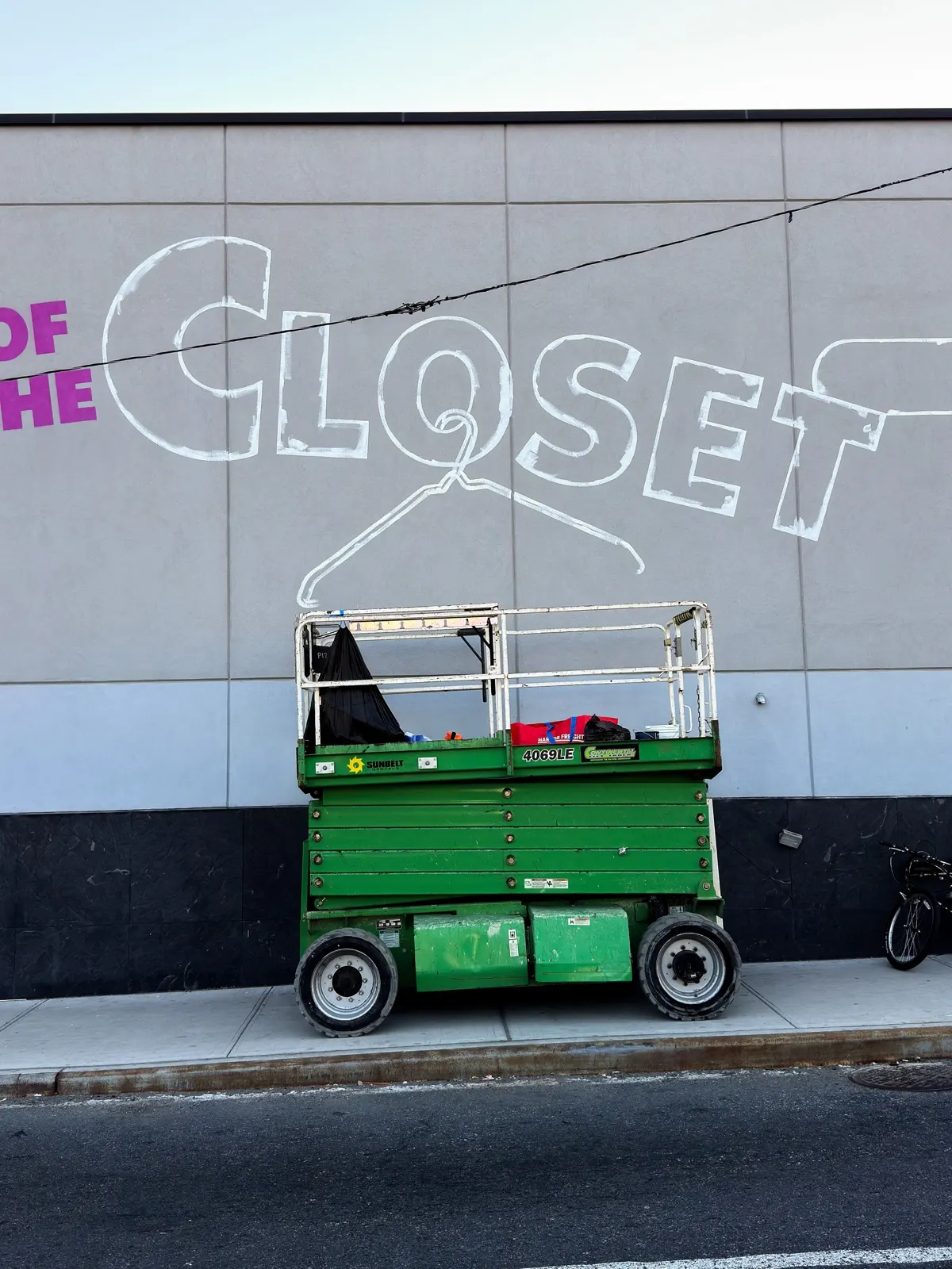 Green rough-terrain scissor lift below the outlined Out of the Closet lettering and hanger during mural setup