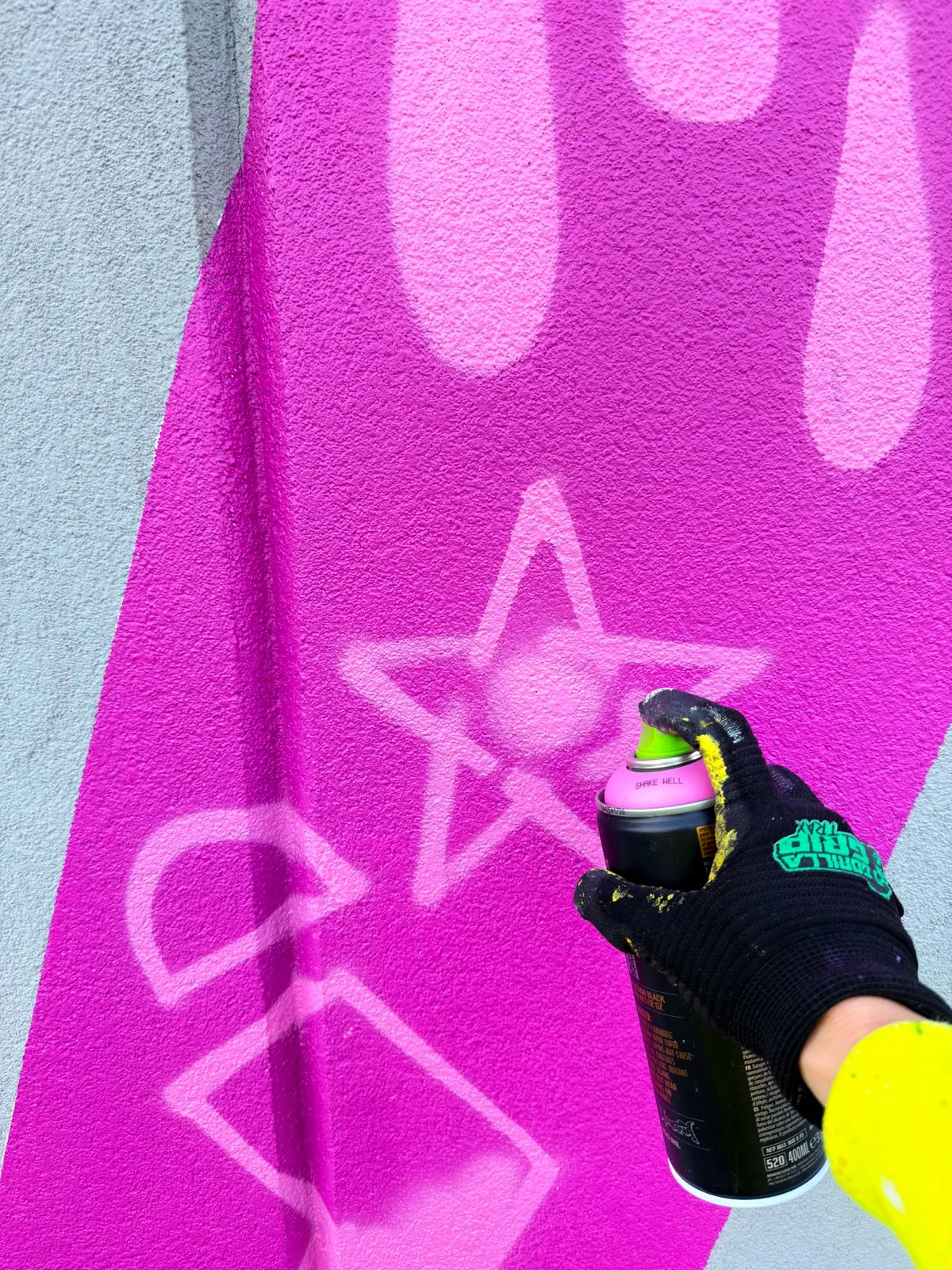 Gloved hand spray-painting a pale pink star and drips inside the bright pink mural lettering