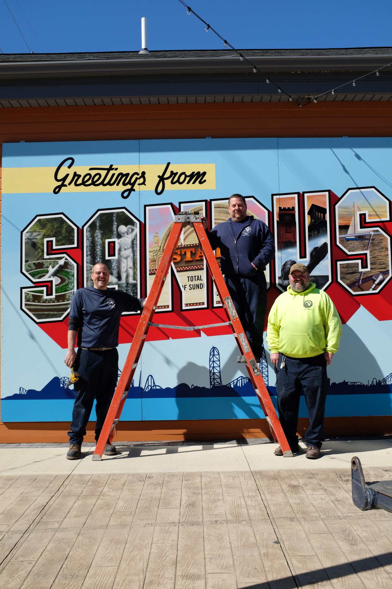 Three installers standing with the completed Greetings from Sandusky mural