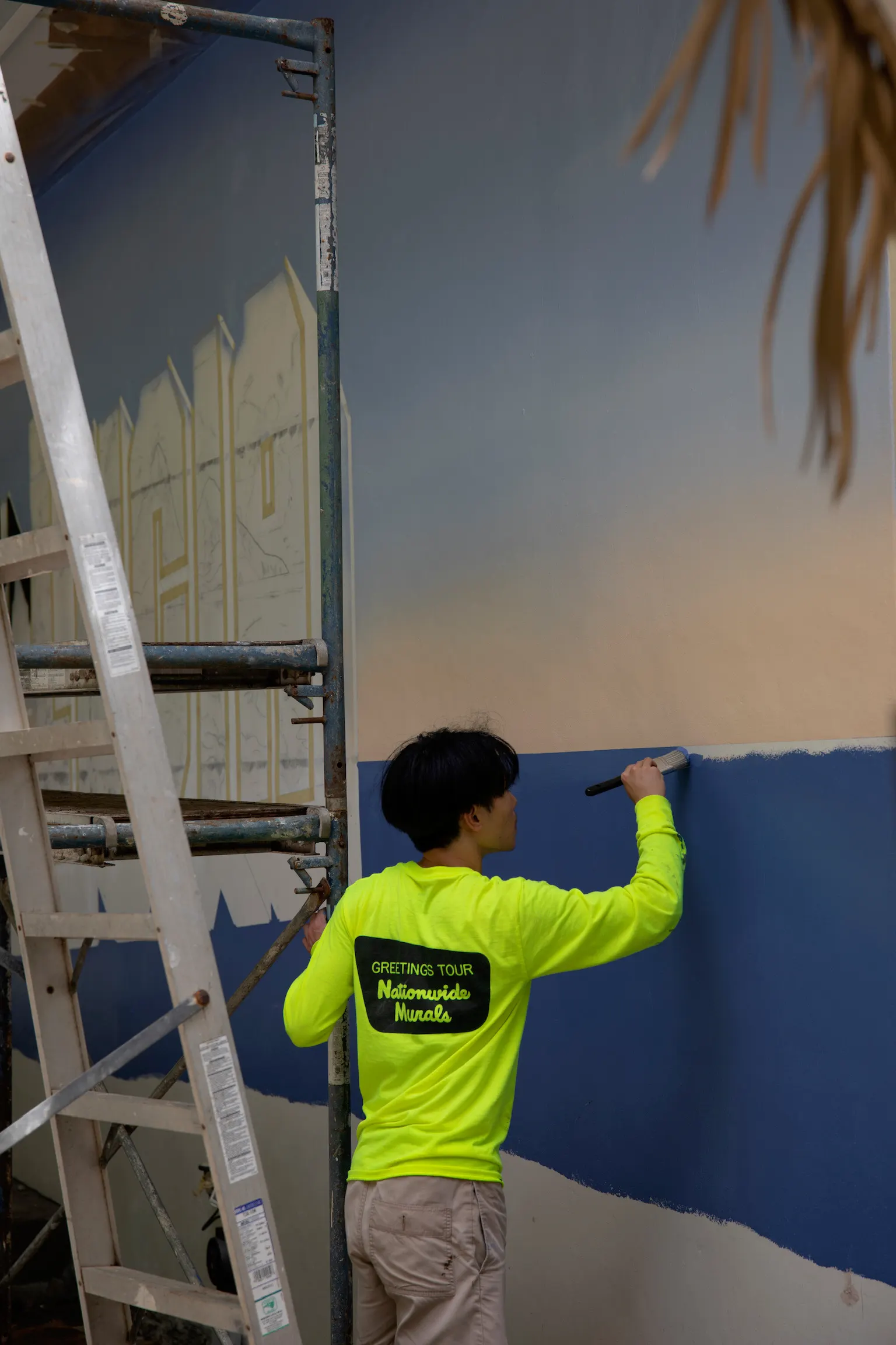 Artist brushing blue paint onto the St. Lucia mural beside ladders and scaffolding