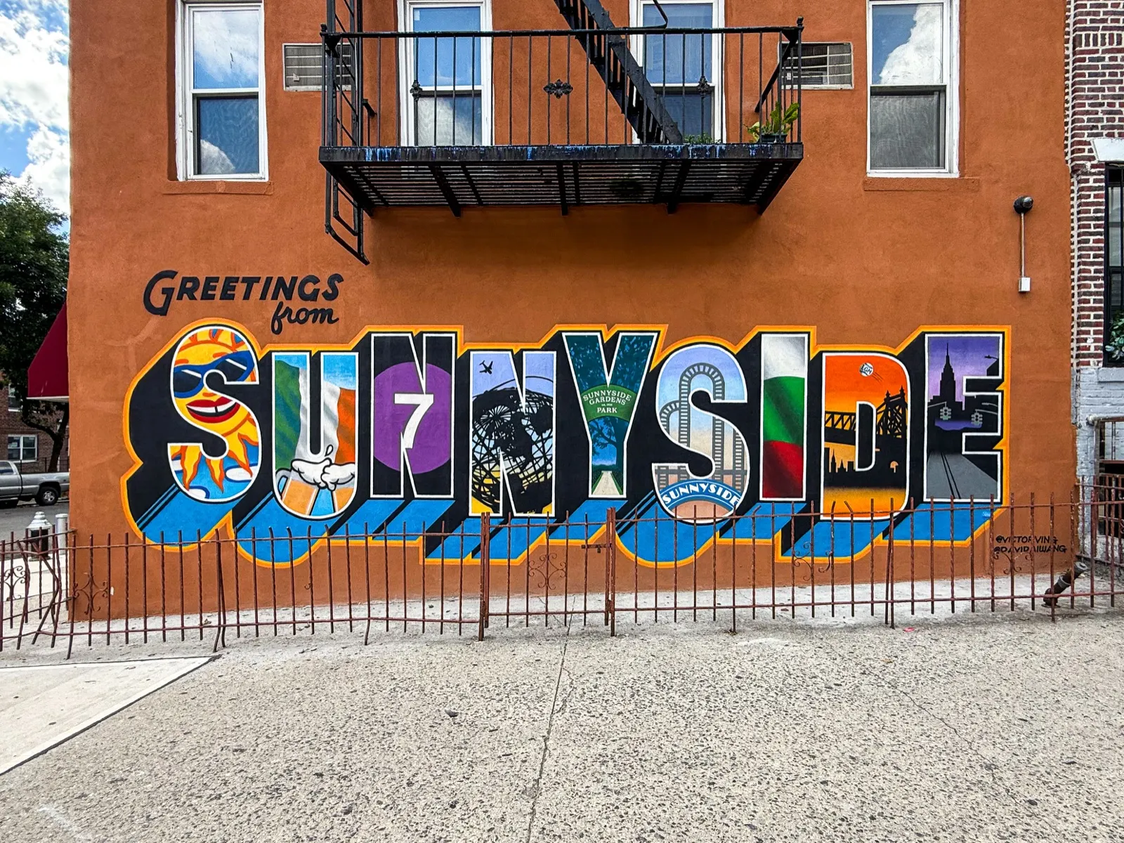 Finished Greetings from Sunnyside mural stretching across a stucco apartment building beneath a fire escape