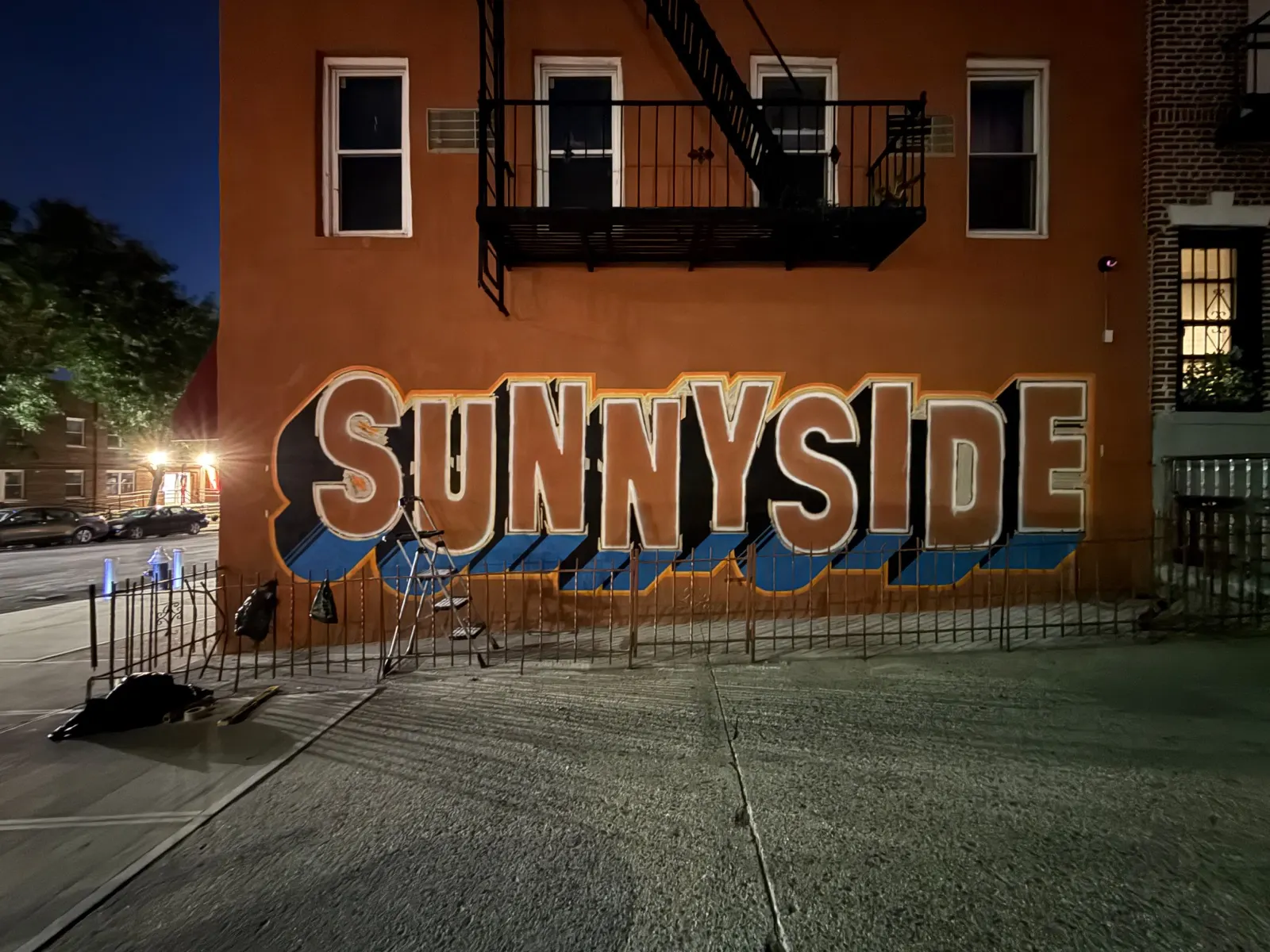Nighttime view of the Sunnyside mural partway through painting beneath the building's fire escape