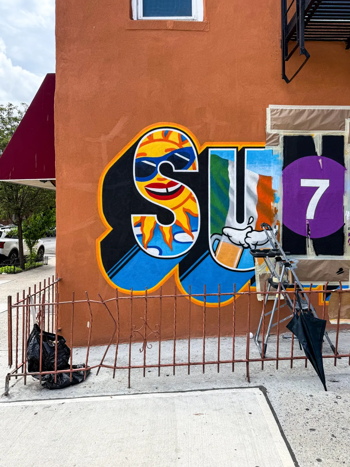 Daylight work-in-progress view of the mural's left side with masking paper, paint, and a stepladder