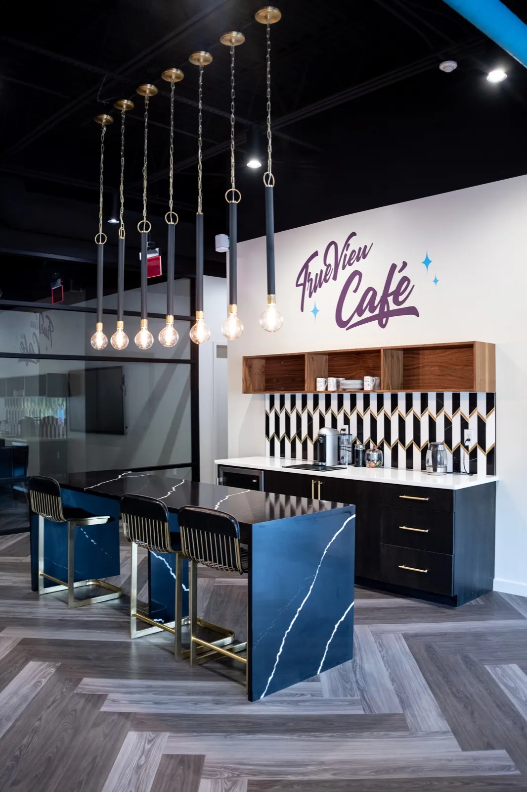 Full office cafe with the TrueView Cafe sign, pendant lights, blue and black island, stools, and coffee counter